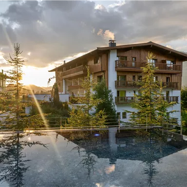 Ein elegantes Hotel mit Holzfassade und Blick auf die Berge. Die Abendsonne strahlt über die Landschaft und spiegelt sich im Pool.