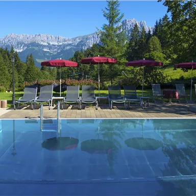Ein klarer Pool mit Liegen und Sonnenschirmen, umgeben von einer schönen, grünen Landschaft. Im Hintergrund sind majestätische Berge zu sehen.