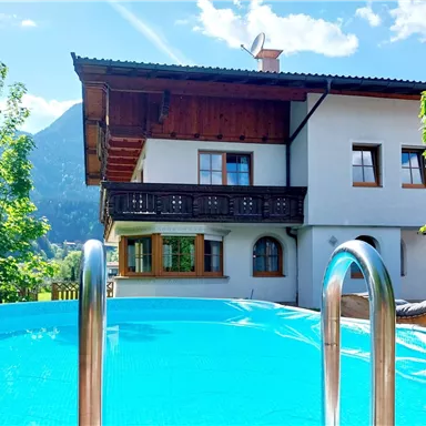 A beautiful house with a pool and a mountain landscape in the background. The sun is shining and there is plenty of greenery around the building.