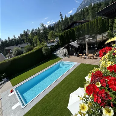 A beautiful garden with a blue pool and colorful flowers. In the background, green trees and mountains can be seen.