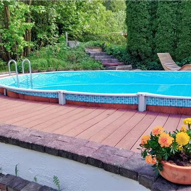 A clean pool with clear water is surrounded by a wooden deck. In the background, there are green trees and a planted corner.