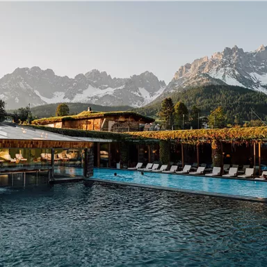 A beautiful pool with loungers, surrounded by mountains. The landscape is partly wooded and snow-capped peaks are visible in the background.