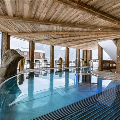 Ein eleganter Wellnessbereich mit einem blauen Pool und Holzverkleidung. Große Fenster bieten einen Blick auf die verschneite Landschaft.