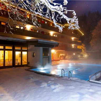 Ein gemütliches Hotel mit einem beheizten Außenpool, umgeben von Schnee. Die Szene wird von einem klaren Nachthimmel und funkelnden Lichtern beleuchtet.