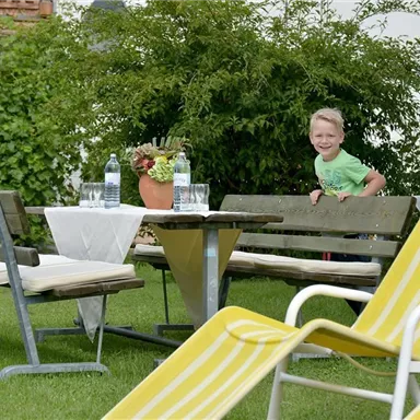 A beautiful garden with a table group and a blooming plant. A happy child stands behind the benches and looks curiously at the camera.
