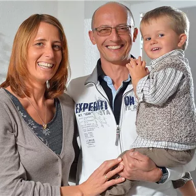Ein glückliches Familienportrait mit einem Paar und ihrem kleinen Kind. Alle lächeln freundlich in einem hellen Raum.