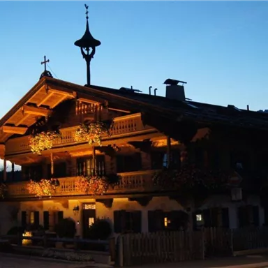 Ein charmantes Holzhaus mit Balkonen, das abends beleuchtet ist. Es ist umgeben von Bergen und hat eine einladende Atmosphäre.