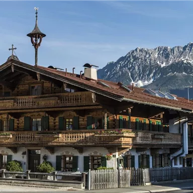 Ein traditionelles alpenländisches Holzhaus mit Balkonen und grünen Fensterläden. Im Hintergrund ragen majestätische Berge unter einem klaren blauen Himmel auf.