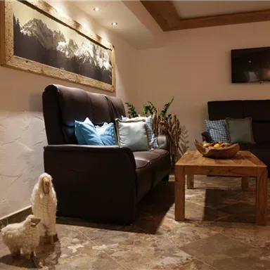 Ein gemütlicher Wohnbereich mit einem schwarzen Sofa und einem Sessel. Der Raum hat eine warme Atmosphäre mit dekorativen Schafen und einem Holztisch.