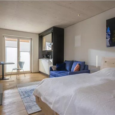 Ein helles Zimmer mit einem bequemen Bett, einem blauen Sofa und einem kleinen Esstisch. Große Fenster bieten viel natürliches Licht.