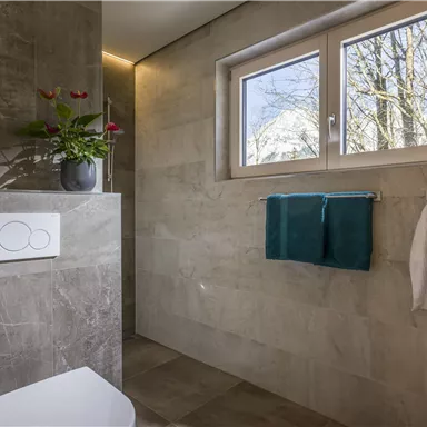 Ein modernes Badezimmer mit einer Toilette, Handtüchern und einem Fenster, das Licht und Ausblick auf Bäume bietet. Die Wände sind in neutralen Farben gehalten.