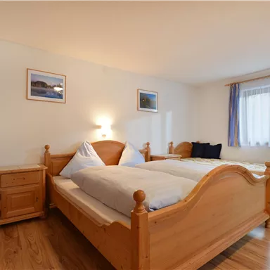 A cozy bedroom with two wooden beds and soft lighting. The walls are adorned with pictures, and a window provides daylight.