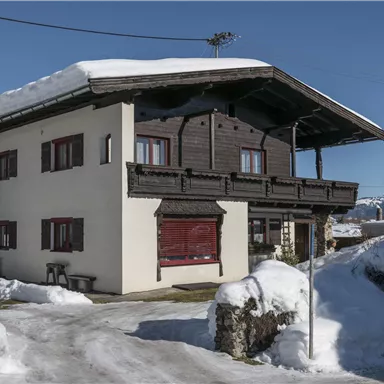 A cozy house in the snow with an upper balcony. The landscape is wintry and the mountains are visible in the background.
