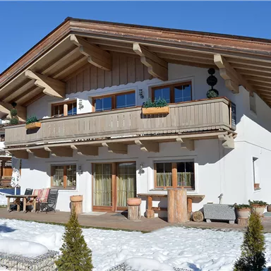 An alpine house with wooden cladding and a large balcony. The exterior is surrounded by snow and small green shrubs.