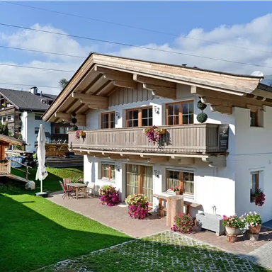 A cozy alpine house with a balcony and flower pots. The garden is green and well-maintained, surrounded by mountains and a clear sky.