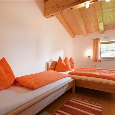 A cozy room with two single beds and orange cushions. The room has wooden ceilings and large windows that provide plenty of light.