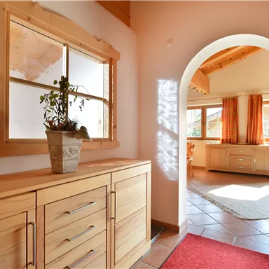 An inviting hallway with a wooden cabinet and a plant on the table. In the background, a bright room with windows and a carpet can be seen.