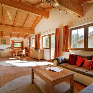 A cozy living room with a wooden beam ceiling and large windows. There is a comfortable sofa, a table, and an open access to the kitchen.