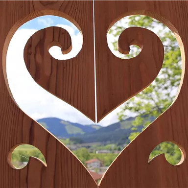 A heart-shaped wooden window overlooking a green landscape with mountains in the background. The sky is blue with clouds.