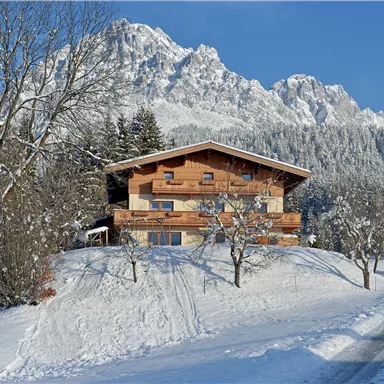 Ein gemütliches Holzhaus in einer verschneiten Winterlandschaft. Im Hintergrund erheben sich majestätische Berge unter einem klaren blauen Himmel.
