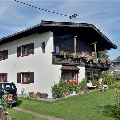 Ein gemütliches Haus mit Balkon und Blumen im Vorgarten. Umgeben von grüner Natur und Bergkulisse.