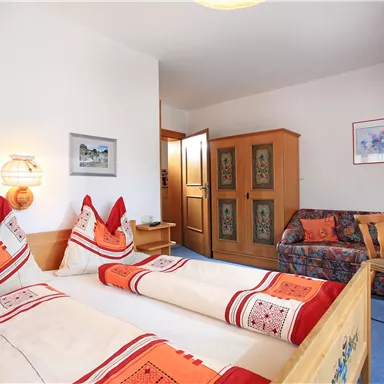 Ein gemütliches Hotelzimmer mit zwei Betten und rot-orangefarbenen Dekokissen. Es gibt einen kleinen Tisch und eine Couch sowie einen Fernseher an der Wand.