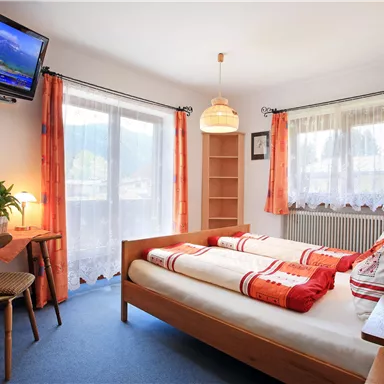 Ein gemütliches Zimmer mit einem Doppelbett und warmen Farben. Es gibt einen Fernseher, einen Tisch und große Fenster mit Vorhängen.