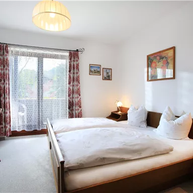 Ein gemütliches Schlafzimmer mit zwei Betten und einem Tisch. Durch das Fenster fällt Tageslicht hinein und es gibt dekorative Bilder an den Wänden.