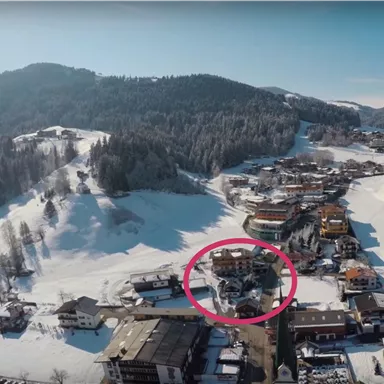 Eine schneebedeckte Landschaft mit einem kleinen Dorf im Vordergrund. Im Hintergrund sind bewaldete Hügel und ein klarer Himmel zu sehen.