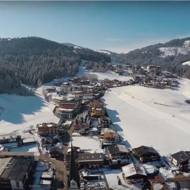 Eine malerische Berglandschaft mit verschneiten Hügeln und einem kleinen Skidorf. Die Sonne scheint über die Winterlandschaft und die Bäume sind mit Schnee bedeckt.