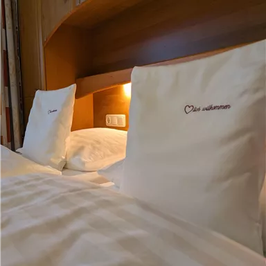 Ein gemütliches Bett mit weißen Kissen und einer sanften Beleuchtung. Die Kissen haben eine entzückende Bestickung.