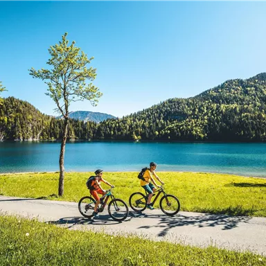 Two children ride their bicycles past a clear lake. The landscape is green and the mountains are visible in the background.