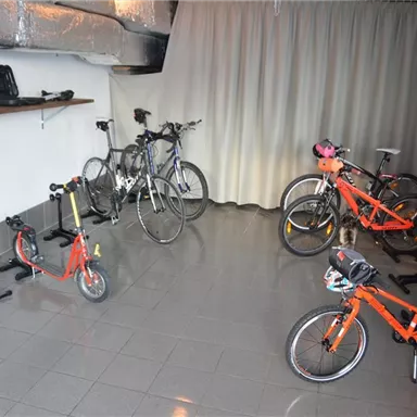 Ein Raum mit mehreren Fahrrädern und einem Kinderroller. Die Wände sind schlicht, und es gibt einen Vorhang sowie einige Regale.