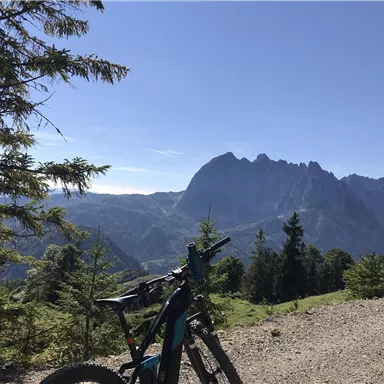 Ein Mountainbike steht auf einem Schotterweg mit Blick auf die Berge. Der Himmel ist klar und die Landschaft ist grün und hügelig.