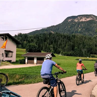 Radfahrer fahren auf einem Weg in einer malerischen ländlichen Umgebung. Im Hintergrund sind grüne Wiesen und Berge zu sehen.