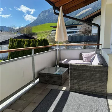 Ein Balkon mit einem gemütlichen Lounge-Bereich und einem Sonnenschirm. Im Hintergrund sind grüne Wiesen und Berge zu sehen.