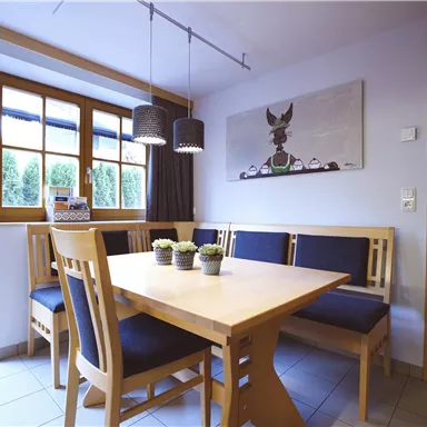 A bright dining area with a wooden table and cozy benches. Large windows let in plenty of daylight.