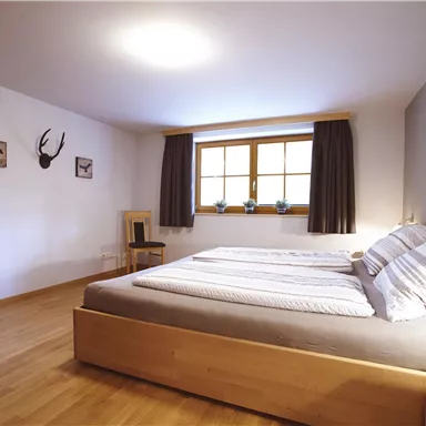 A modern bedroom with a double bed and wooden floor. The windows provide natural light and a cozy atmosphere.