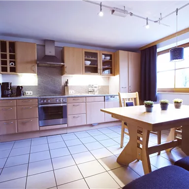 A modern kitchen with wooden cabinets and a dining table. It is bright and spacious, featuring large windows and a cozy ambiance.