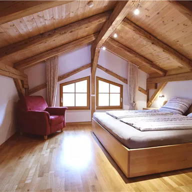 A cozy attic room with a large bed and a red armchair. The wooden ceilings and the floor give the room a warm atmosphere.