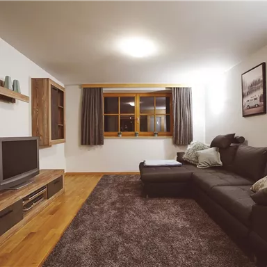 A modern living room with a comfortable black sofa and a television. Large windows provide plenty of daylight, and the walls are decorated with pictures.