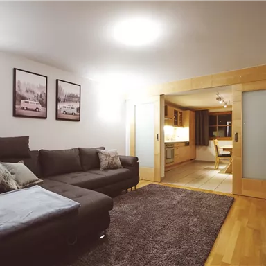 A modern living room with a large, comfortable sofa and a rug. In the background, the kitchen is visible, complemented by some stylish wall art.