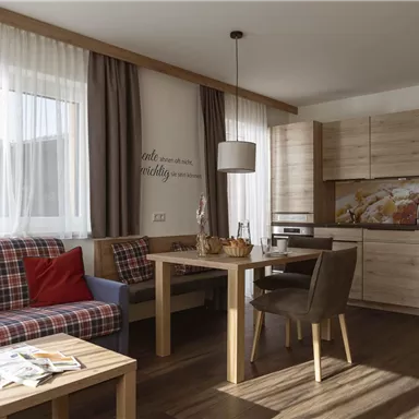 Ein helles Wohnzimmer mit einer gemütlichen Couch, einem Tisch und einer modernen Küchenzeile. Große Fenster sorgen für viel Licht und eine einladende Atmosphäre.