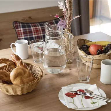 Ein schön gedeckter Tisch mit frischen Brötchen, Obst und Gläsern. Im Hintergrund steht eine Karaffe mit Wasser und eine Blume.
