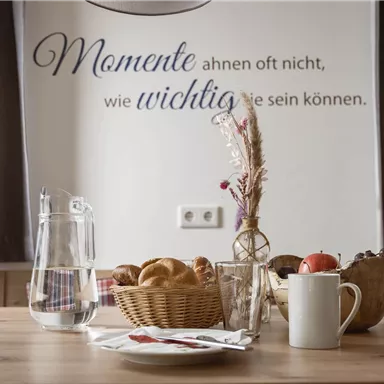 Ein gemütlicher Tisch mit einem Obstkorb, Brötchen und einer Kaffeetasse. Im Hintergrund hängt ein Zitat an der Wand.