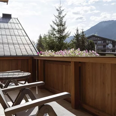 Ein gemütlicher Balkon mit Holzgeländer und zwei Liegen. Im Hintergrund sind Berge und ein blauer Himmel zu sehen.