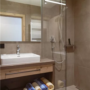 Ein modernes Badezimmer mit einer begehbaren Dusche und einem Waschbecken. Handtücher sind ordentlich auf einem Holzregal verstaut.