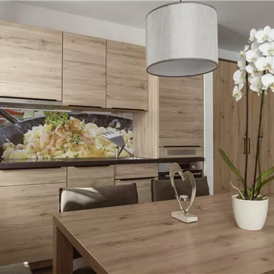 Eine moderne Küche aus Holz mit einem Esstisch und schönen Orchideen. Im Hintergrund sieht man eine Wandverkleidung mit einer bunten Darstellung von Lebensmitteln.