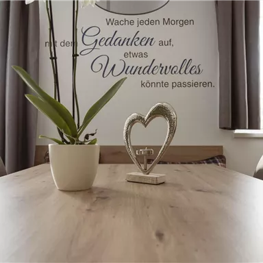 Ein einladender Tisch mit einer Pflanze und einer dekorativen Herzfigur. Im Hintergrund steht ein Spruch an der Wand, der positive Gedanken fördert.