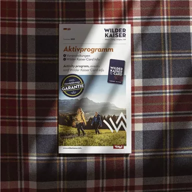 Ein Informationsheft zum Aktivprogramm im Bereich Wilder Kaiser liegt auf einem traditionellen Karomuster. Es zeigt Aktivitäten in der Natur und bietet praktische Informationen für Besucher.